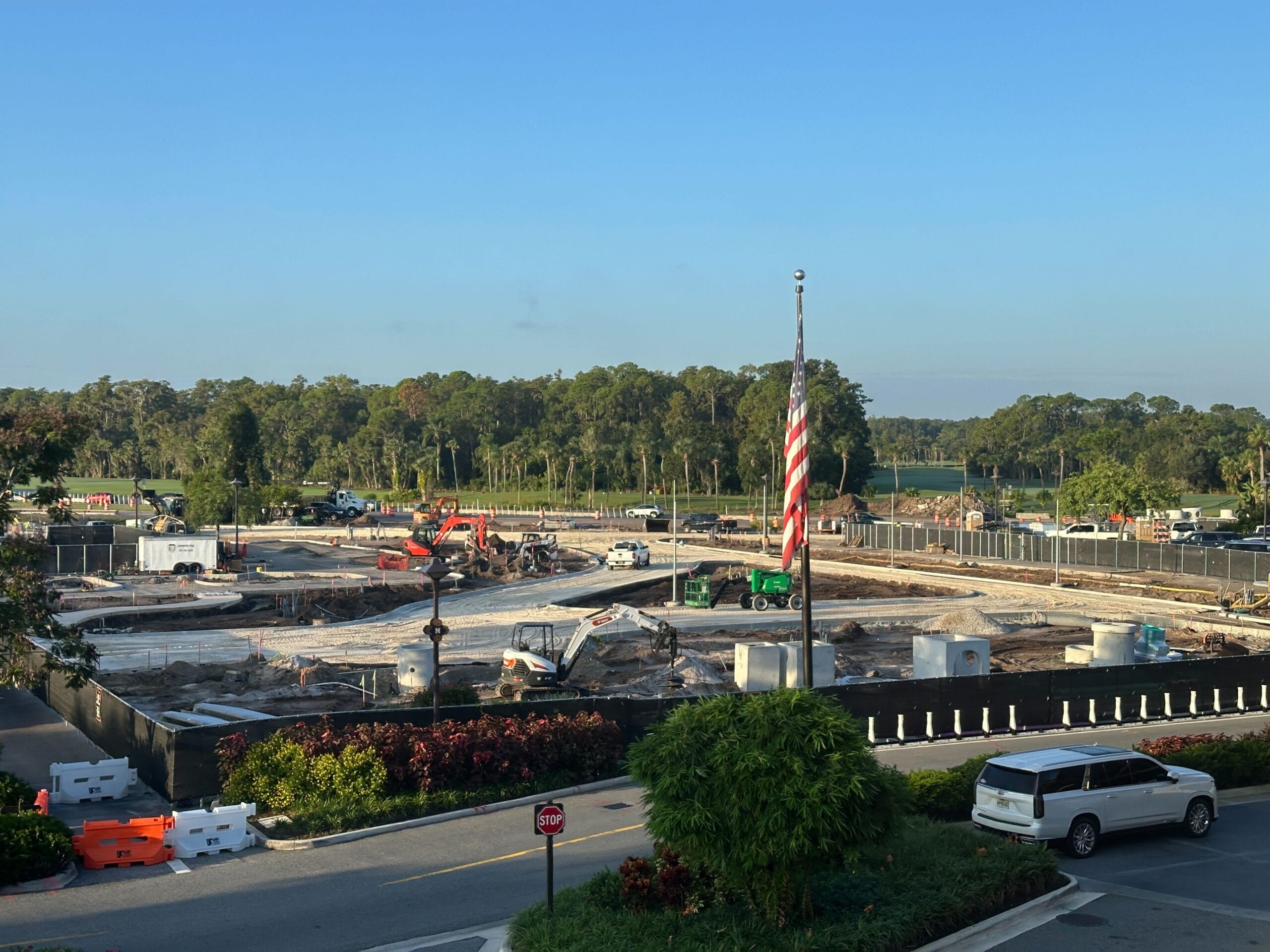 Construction site with vehicles and workers, American flag in foreground, trees and cars in the background under a clear sky. Construction site with vehicles and workers, American flag in foreground, trees and cars in the background under a clear sky.