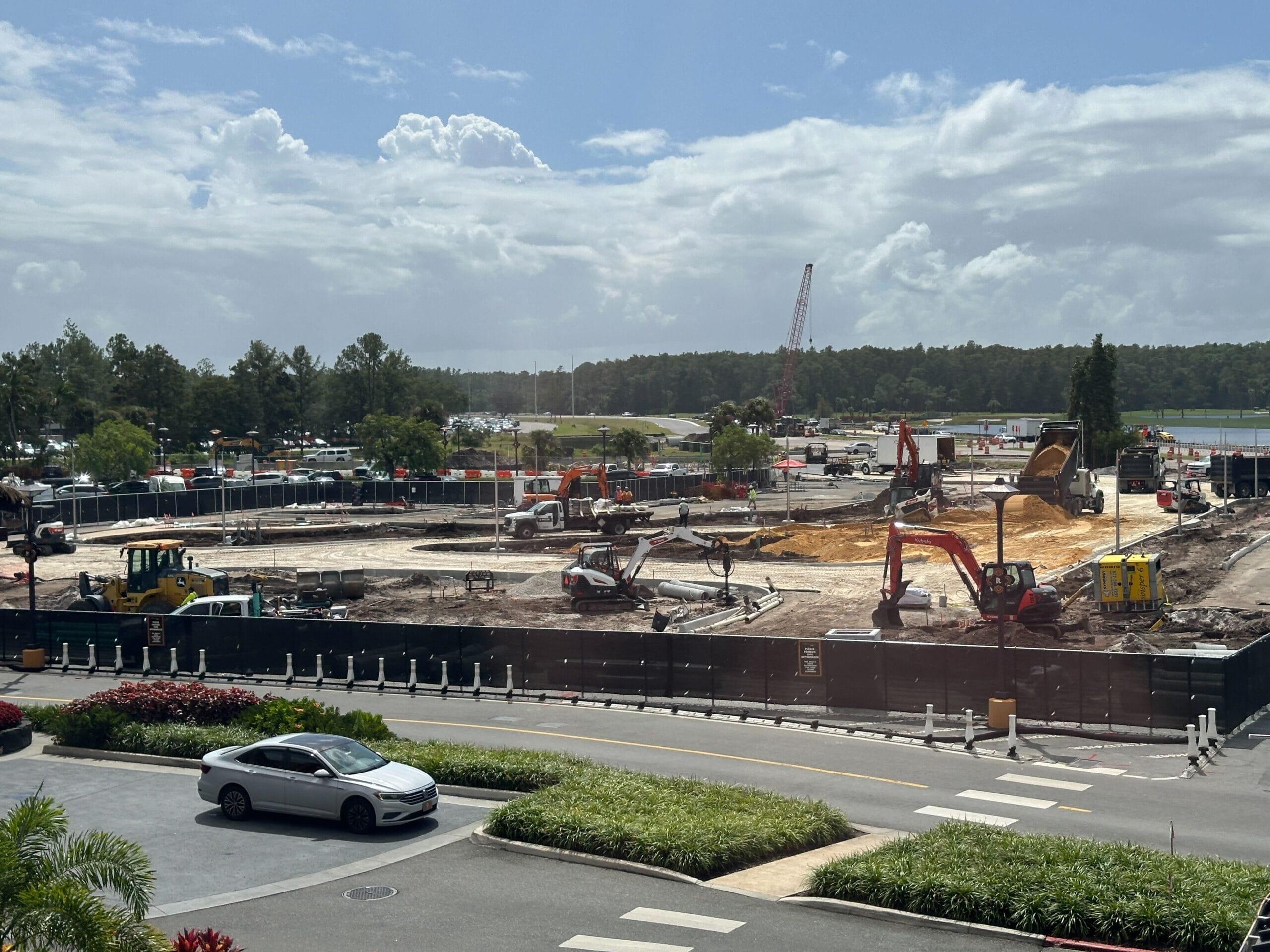 Construction site with various excavators and vehicles, bordered by fencing, trees, and a parked white car in front. Construction site with various excavators and vehicles, bordered by fencing, trees, and a parked white car in front.