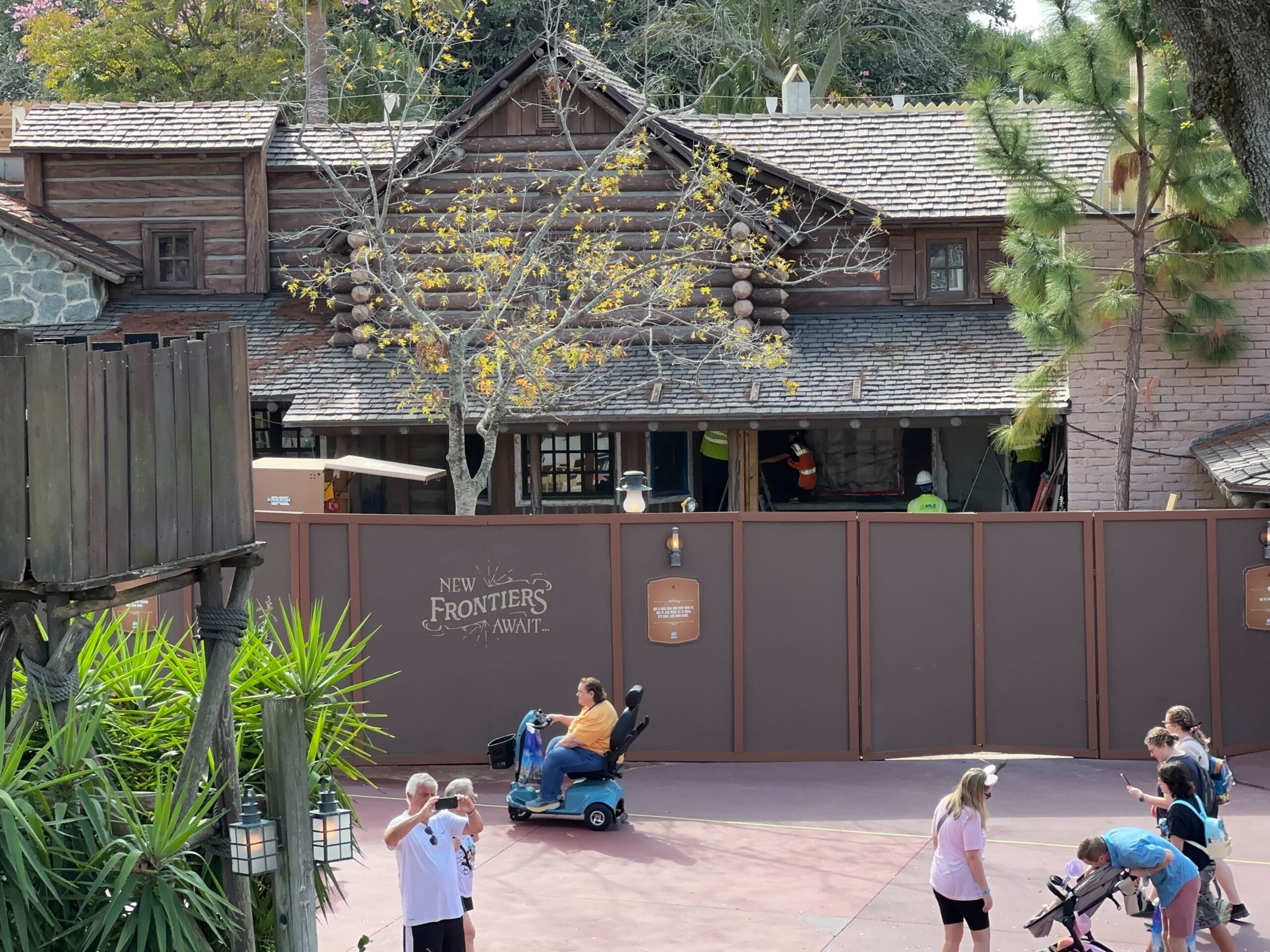 Construction site in front of a log cabin-style building with a sign reading New Frontiers Await. People are walking and a person in a mobility scooter passes by. Trees and greenery are visible.