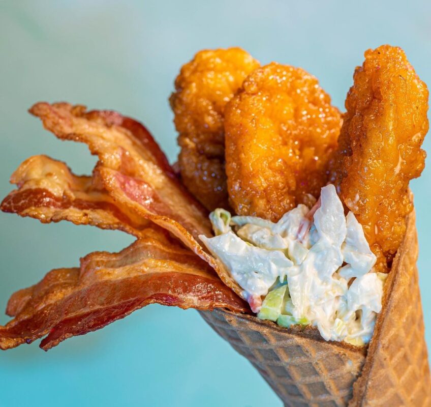 A waffle cone filled with fried chicken strips, strips of bacon, and coleslaw, perfect for enjoying at Typhoon Lagoon's Disney H2O Glow After Hours against a light blue background.'s Disney H2O Glow After Hours against a light blue background.