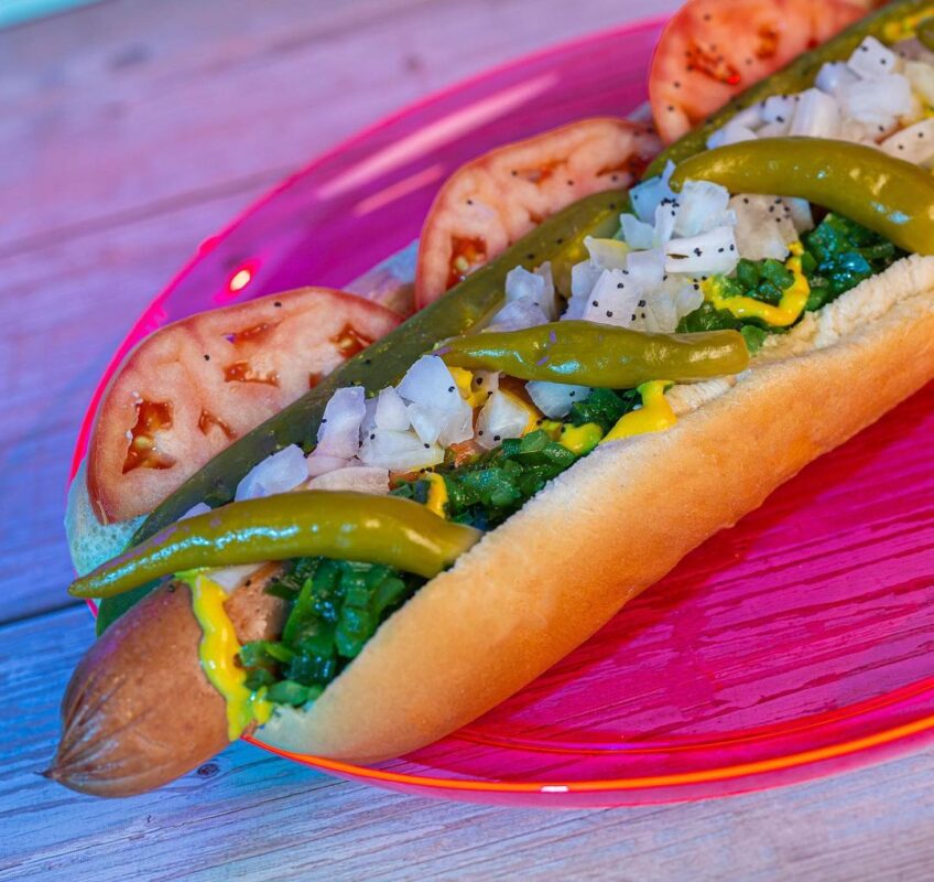A hot dog with toppings including tomato slices, pickle spear, diced onions, relish, mustard, and sport peppers served on a pink plate—a delightful addition to the new menu items at Typhoon Lagoon.