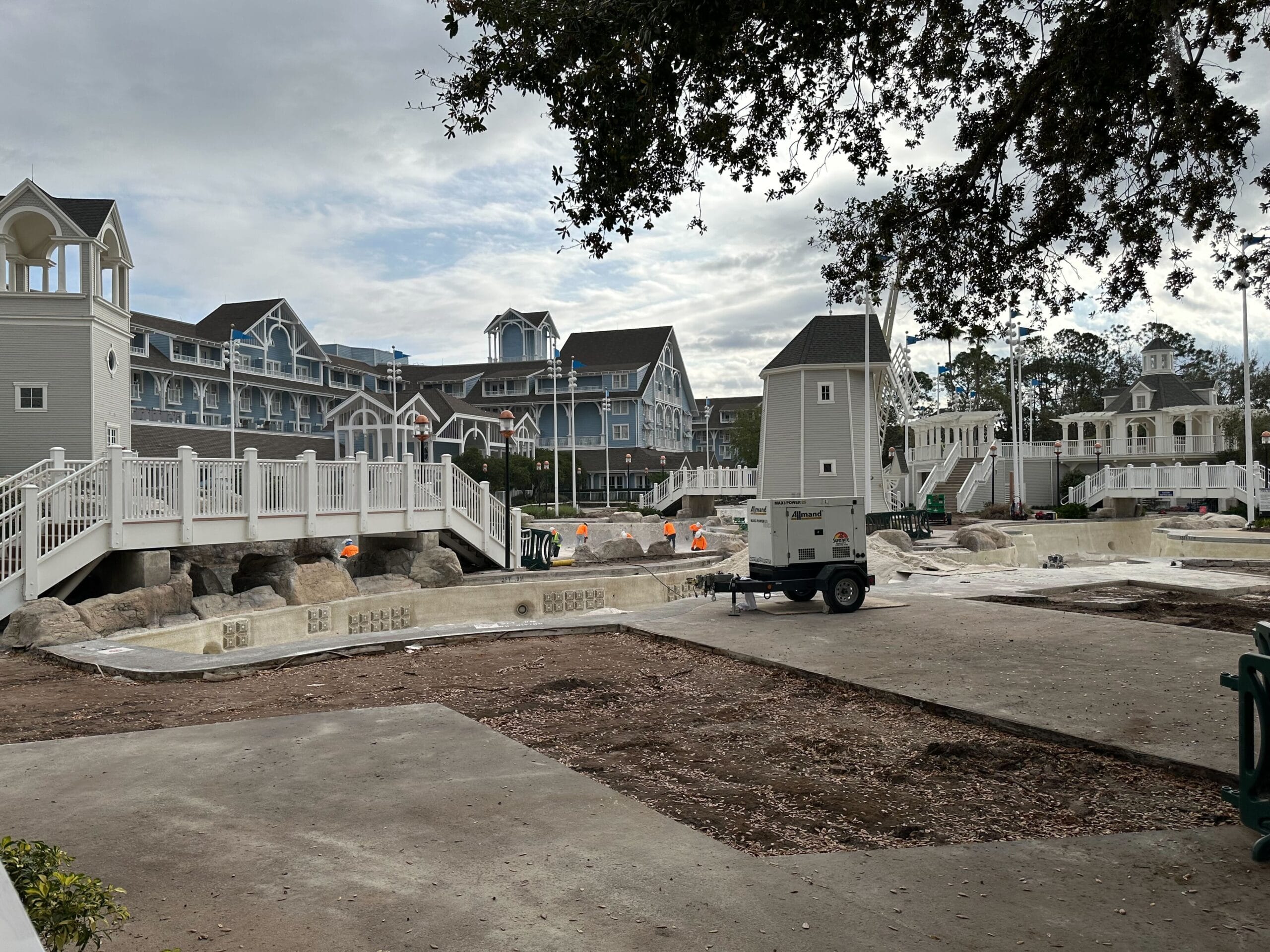 Hotel pool area under construction with empty pool, construction equipment, and hotel buildings in the background. Overcast sky above.