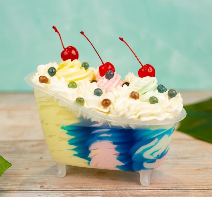 A colorful, bath-shaped dessert with whipped cream, cherries, and candy decorations sits on a wooden surface, inspired by the new menu items at Typhoon Lagoon's Disney H2O Glow.