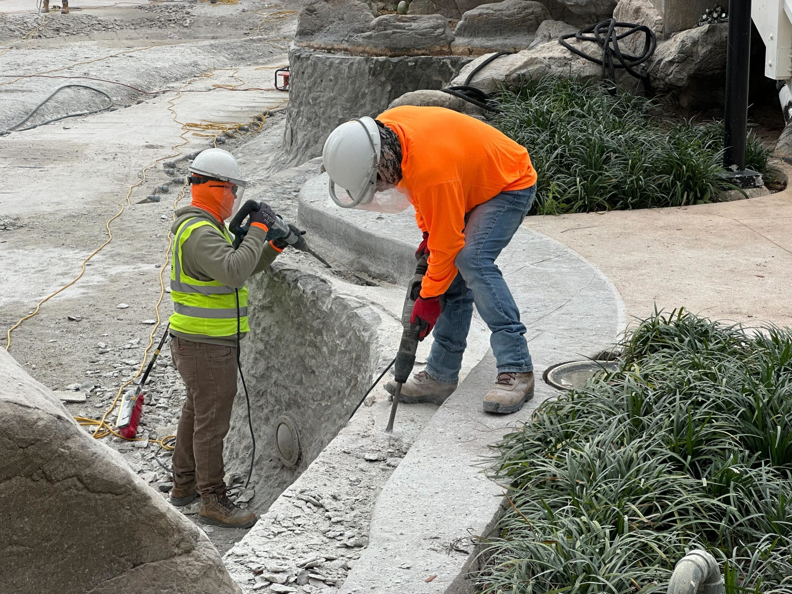 Two construction workers wearing safety gear are using tools to work on a curved concrete structure surrounded by plants.