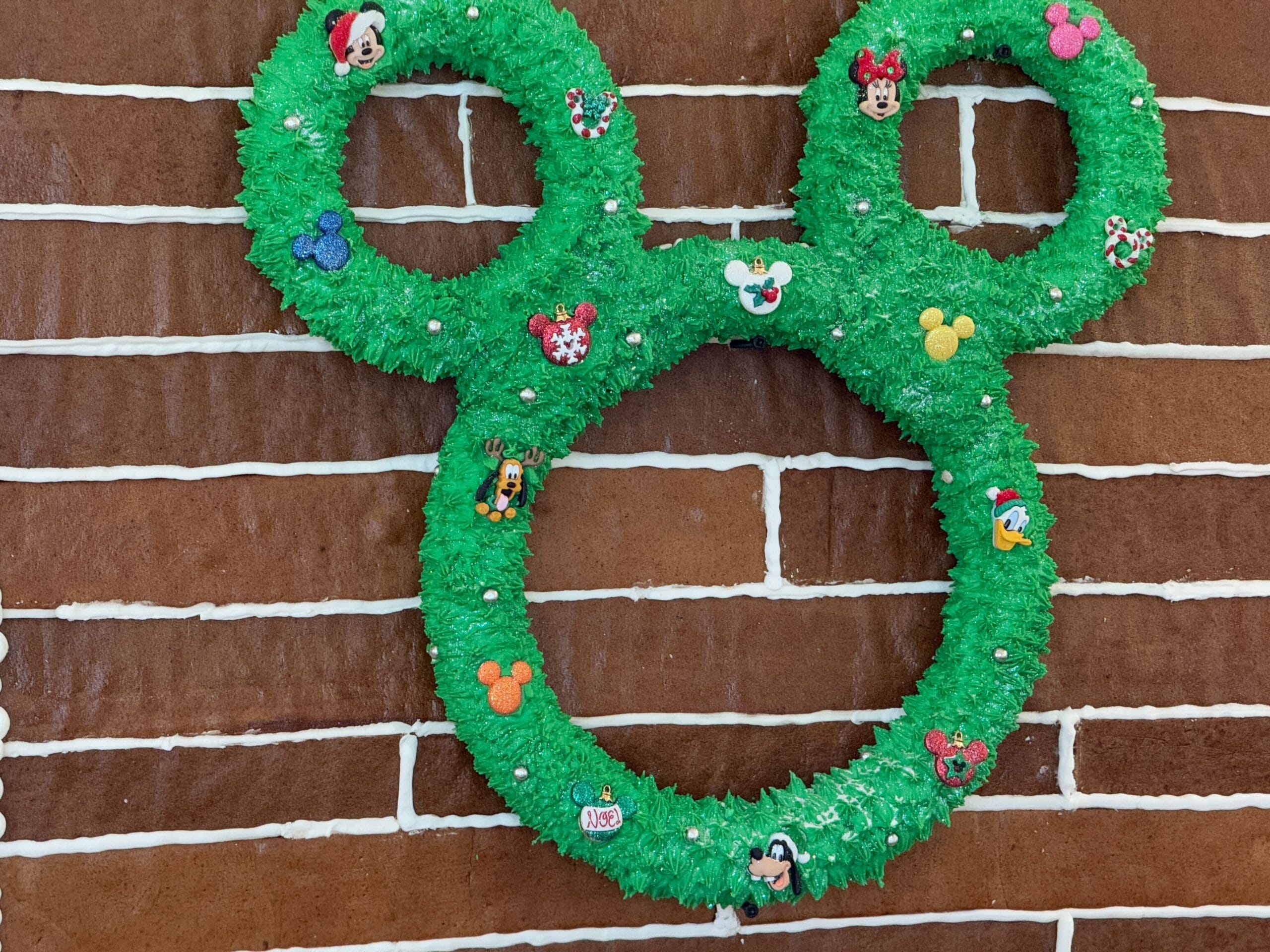 A wreath shaped like a mouse head is adorned with various cartoon character decorations, displayed on a brick-patterned wall.