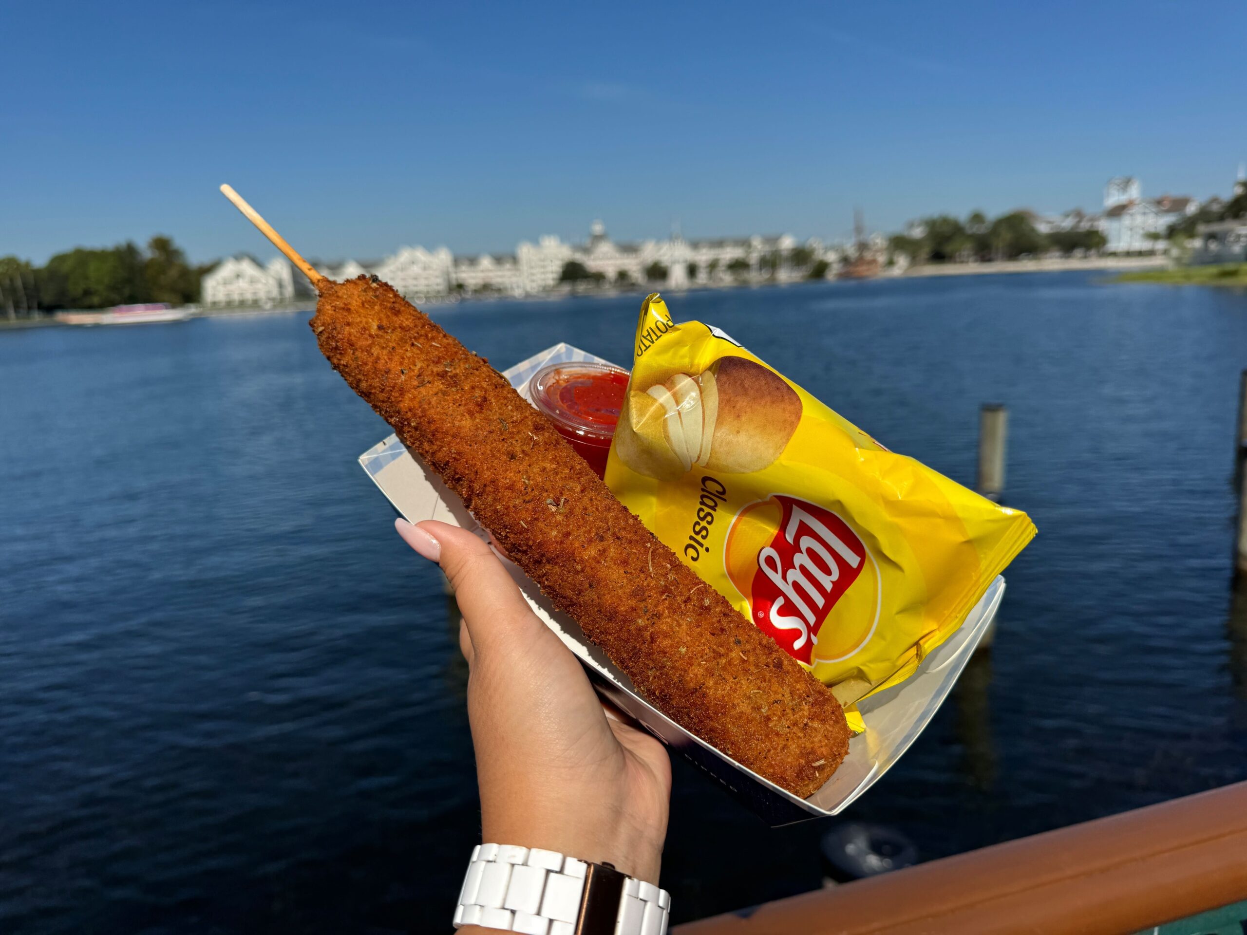 A hand holds a deep-fried corn dog on a stick and a bag of Lay's Classic chips against a scenic waterfront backdrop.'s Classic chips against a scenic waterfront backdrop. A hand holds a deep-fried corn dog on a stick and a bag of Lay's Classic chips against a scenic waterfront backdrop.'s Classic chips against a scenic waterfront backdrop.