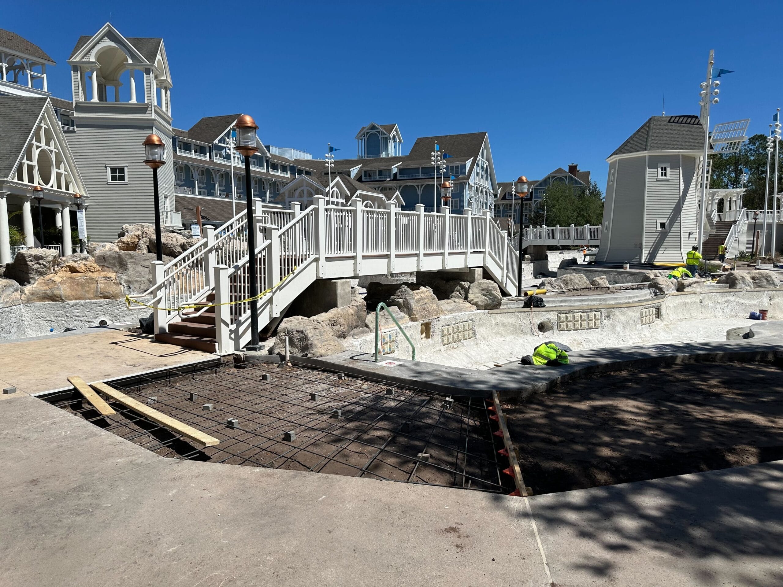 At Disney's Yacht Club, the construction site buzzes with activity near the Stormalong Bay Pool. Workers are busy near a bridge as buildings with gabled roofs stand gracefully in the background under a clear blue sky. At Disney's Yacht Club, the construction site buzzes with activity near the Stormalong Bay Pool. Workers are busy near a bridge as buildings with gabled roofs stand gracefully in the background under a clear blue sky.