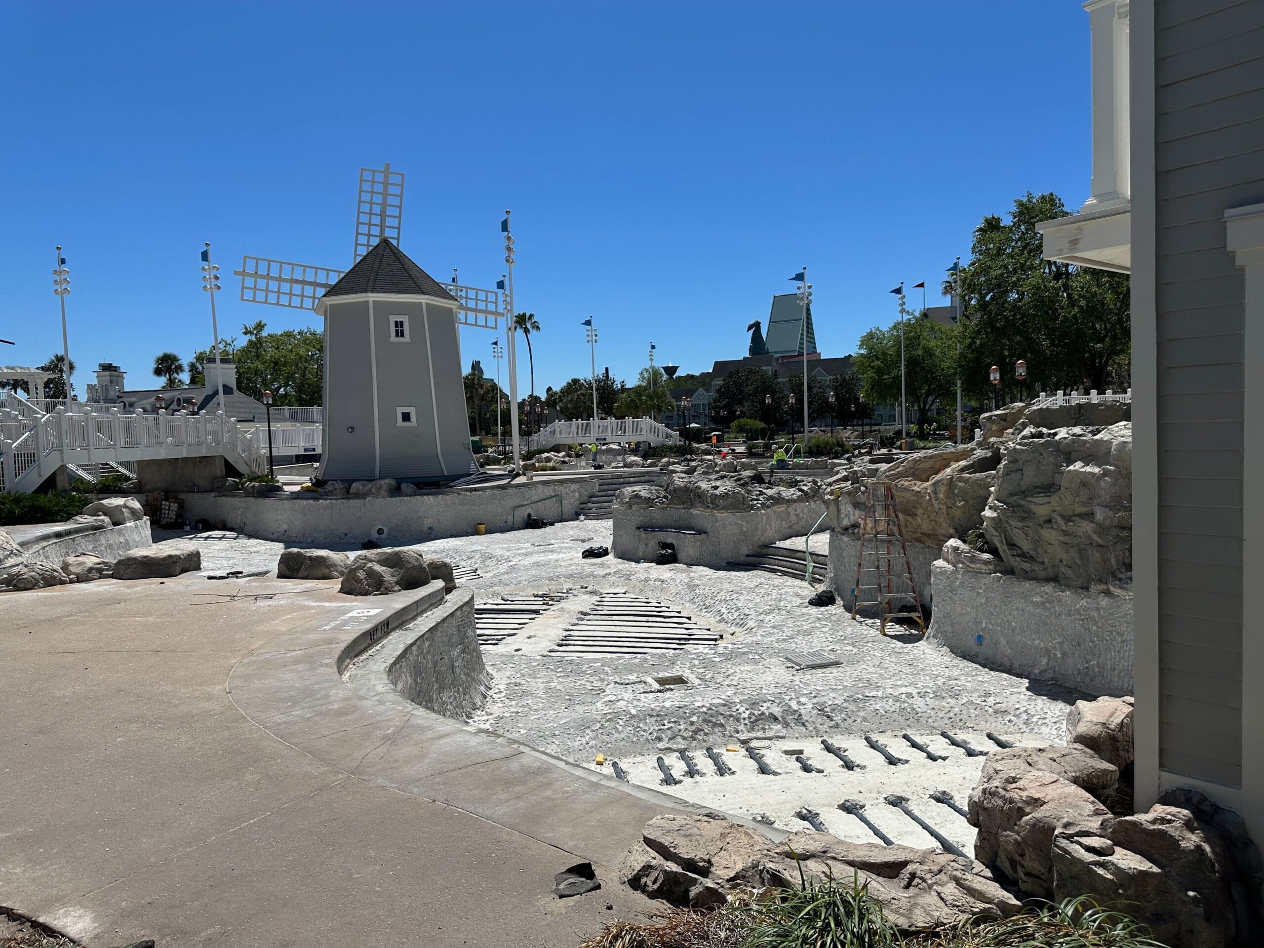 The construction site at Disney's Yacht Club features an empty Stormalong Bay Pool, a ladder, and a windmill structure under a clear blue sky. Rocks and unfinished surfaces are visible in the fenced-off area. The construction site at Disney's Yacht Club features an empty Stormalong Bay Pool, a ladder, and a windmill structure under a clear blue sky. Rocks and unfinished surfaces are visible in the fenced-off area.