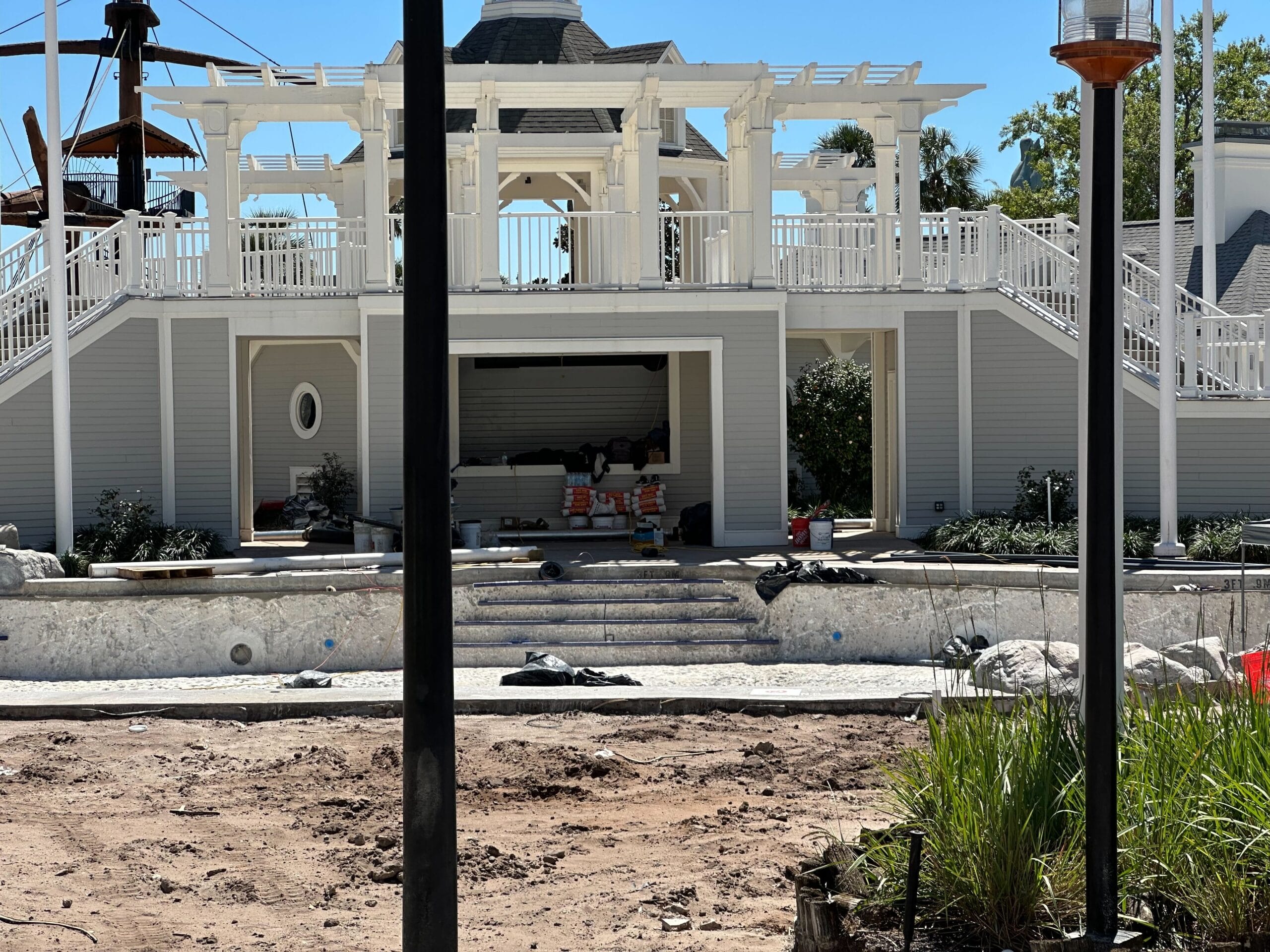 A building under construction stands beside the Stormalong Bay Pool, with a foreground of dirt and construction materials. Steps and a white pergola lead to the entrance of Disney's Yacht Club, seamlessly blending luxury with progress. A building under construction stands beside the Stormalong Bay Pool, with a foreground of dirt and construction materials. Steps and a white pergola lead to the entrance of Disney's Yacht Club, seamlessly blending luxury with progress.