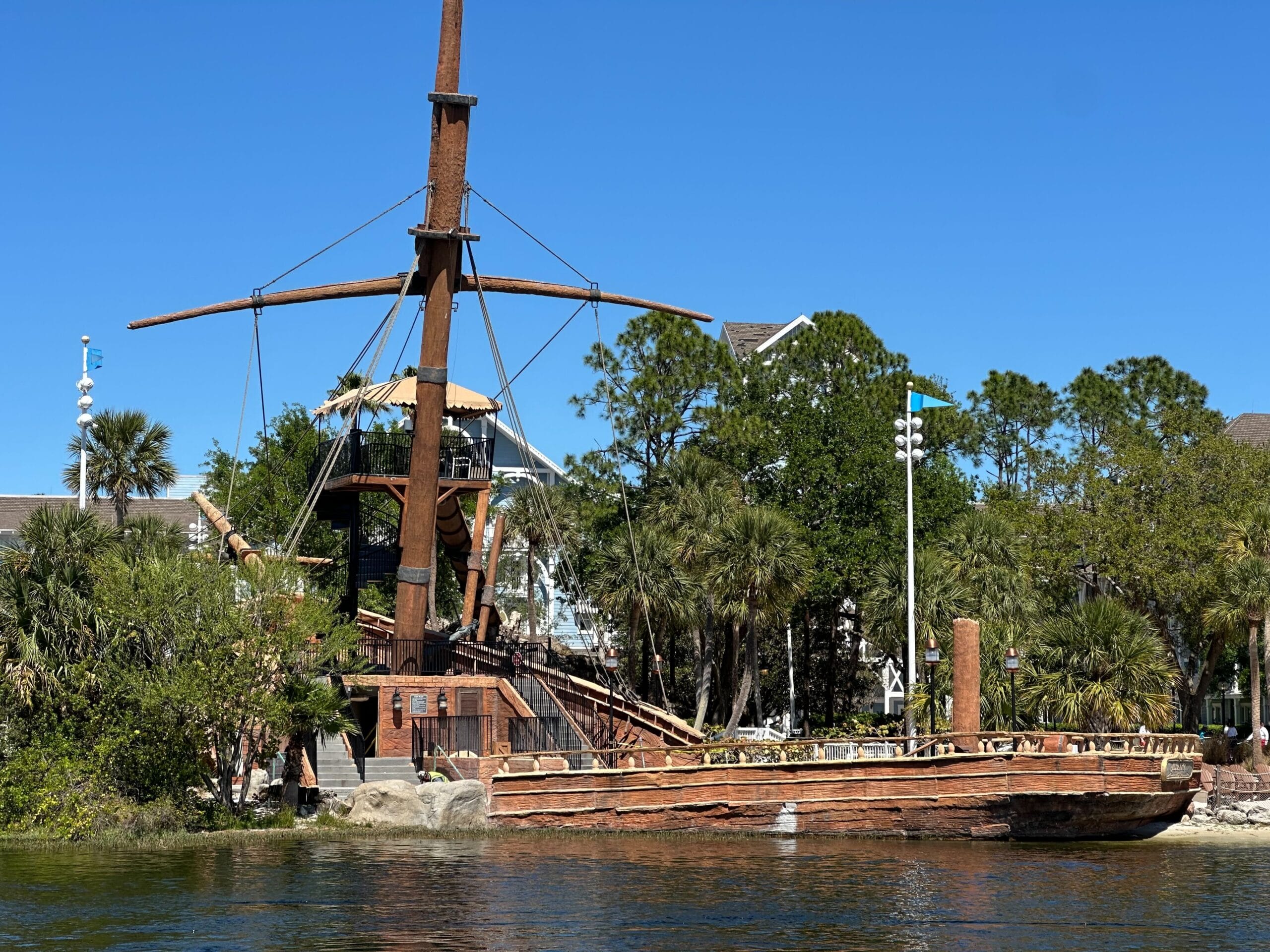 A wooden pirate ship with masts is docked by a lush, tree-lined shore under a clear blue sky, reminiscent of the enchanting ambiance at Disney’s Yacht Club. A wooden pirate ship with masts is docked by a lush, tree-lined shore under a clear blue sky, reminiscent of the enchanting ambiance at Disney’s Yacht Club.