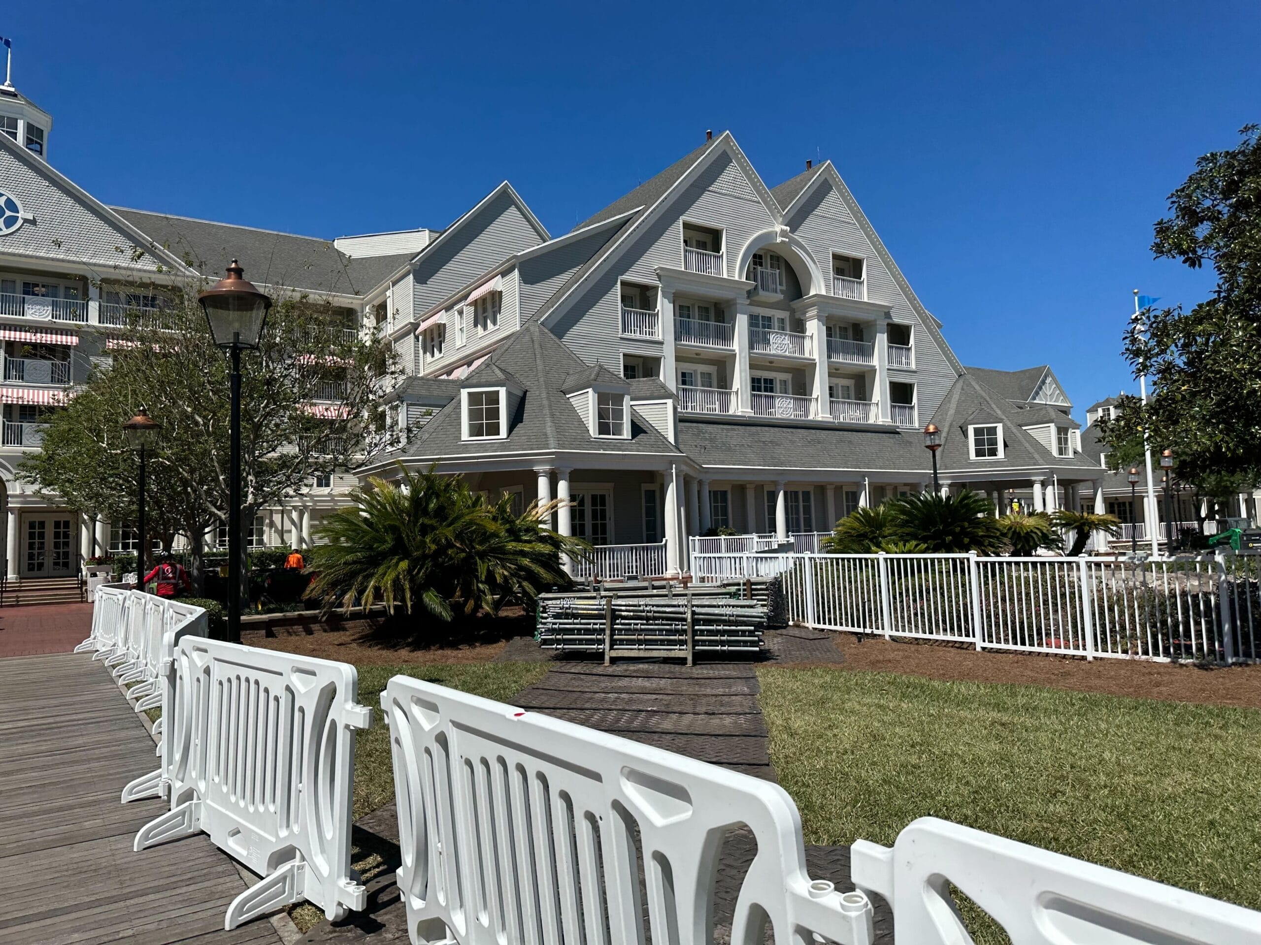 A large, light-gray building reminiscent of Disney’s Yacht Club stands under a clear blue sky. White barriers, hinting at ongoing construction, line a wooden walkway in the foreground, with lush greenery and a tree completing the serene scene. A large, light-gray building reminiscent of Disney’s Yacht Club stands under a clear blue sky. White barriers, hinting at ongoing construction, line a wooden walkway in the foreground, with lush greenery and a tree completing the serene scene.