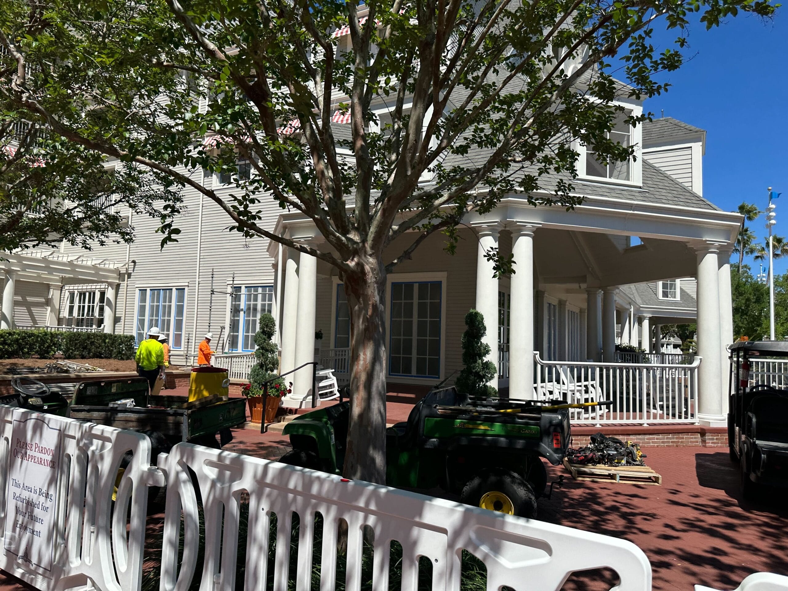 A two-story house with columns and a porch, reminiscent of Disney’s Yacht Club, is surrounded by trees. Construction workers and equipment are busy in front, with a white barricade adding to the scene's dynamic energy. A two-story house with columns and a porch, reminiscent of Disney’s Yacht Club, is surrounded by trees. Construction workers and equipment are busy in front, with a white barricade adding to the scene's dynamic energy.