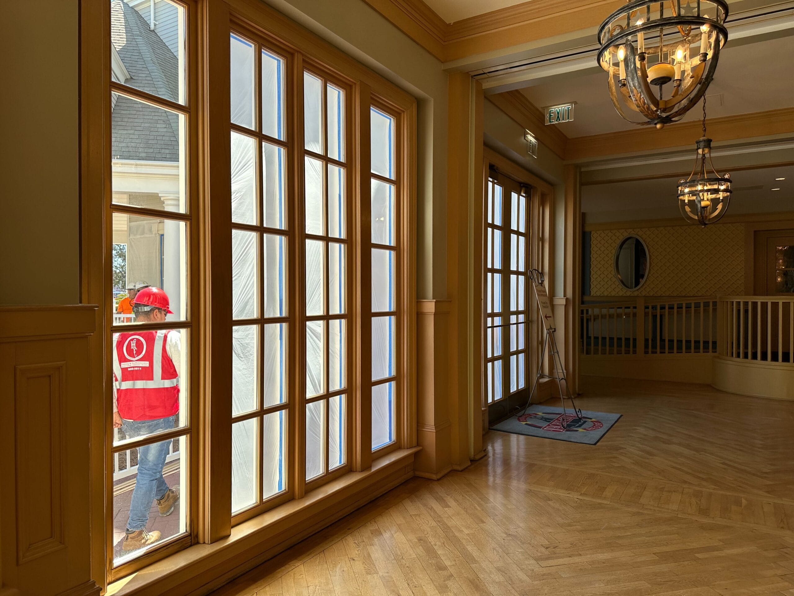 A person in a red safety vest and hard hat walks outside a window, possibly part of the construction work at Disney’s Yacht Club. Indoors, there's wooden flooring, decorative lighting, and a ladder near a door. A person in a red safety vest and hard hat walks outside a window, possibly part of the construction work at Disney’s Yacht Club. Indoors, there's wooden flooring, decorative lighting, and a ladder near a door.